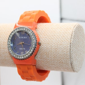 May include: An orange quartz watch with a blue face and a silver bezel encrusted with rhinestones. The watch has the word "AUBURN" and "QUARTZ" printed on the face. The watch is displayed on a beige cylindrical stand.