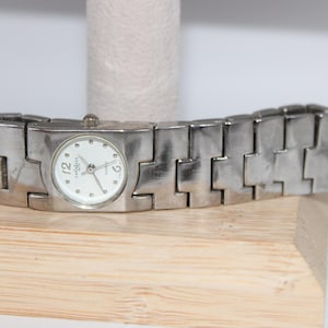 May include: A silver-toned wristwatch with a white face and a rectangular case. The watch features a link-style band and is displayed on a wooden block. The watch face has the brand name and the numbers 12, 6, and 2.