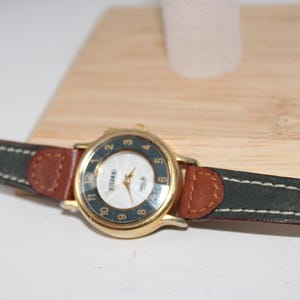 May include: A gold-toned wristwatch with a white face and dark green bezel. The watch has a brown leather strap with dark green and white stitching accents. The watch is displayed on a wooden stand.