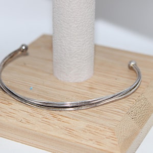 May include: A silver-colored cuff bracelet with a simple, elegant design. The bracelet features a double-banded structure with small, spherical accents at each end. It rests on a light-colored wooden display stand.