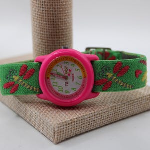 May include: A pink Timex watch with a green fabric strap decorated with embroidered red dragonflies. The watch face has white numerals and hands, with the text "TO FAST" and the Timex logo. The watch is displayed on a small, square, tan fabric-covered block.