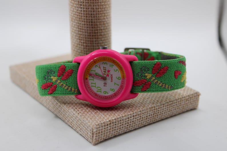 May include: A pink Timex watch with a green fabric strap decorated with embroidered red dragonflies. The watch face has white numerals and hands, with the text "TO FAST" and the Timex logo. The watch is displayed on a small, square, tan fabric-covered block.
