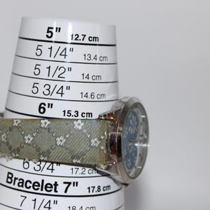 May include: A silver wristwatch with a light blue face and a patterned band, resting on a bracelet sizing tool. The tool displays measurements from 5 inches (12.7 cm) to 7 1/4 inches (18.4 cm).