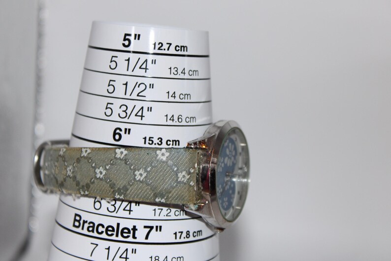 May include: A silver wristwatch with a light blue face and a patterned band, resting on a bracelet sizing tool. The tool displays measurements from 5 inches (12.7 cm) to 7 1/4 inches (18.4 cm).