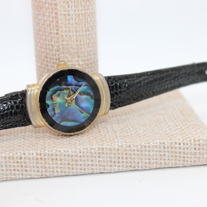 May include: A vintage wristwatch with a gold-tone case and a black leather band. The watch face features a shimmering, iridescent abalone shell design. The watch is displayed on a beige fabric surface.
