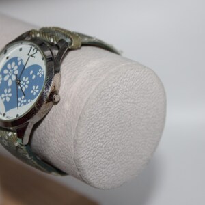May include: Silver-toned wristwatch with a white face featuring a blue heart design filled with white floral patterns. The watch has a patterned band in shades of green and tan.