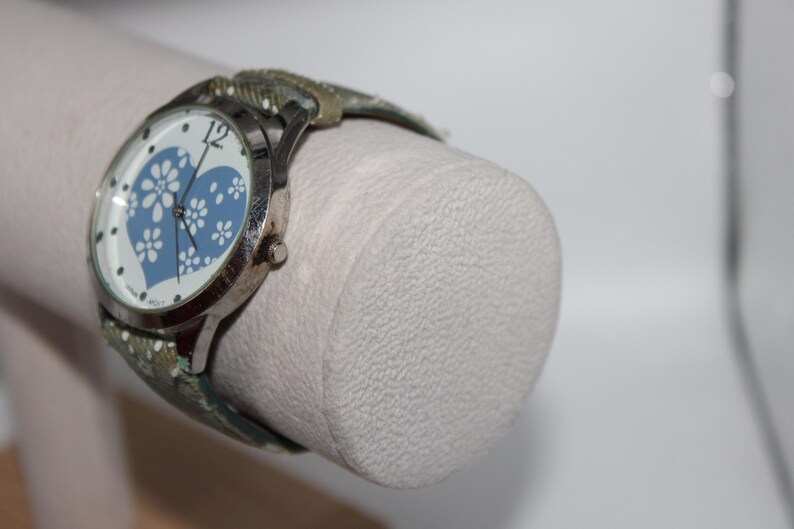May include: Silver-toned wristwatch with a white face featuring a blue heart design filled with white floral patterns. The watch has a patterned band in shades of green and tan.