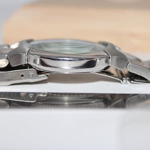 May include: A silver-toned wristwatch with a metallic band. The watch face is visible, and the band is connected with a series of small, linked segments. The watch is displayed on a reflective surface.