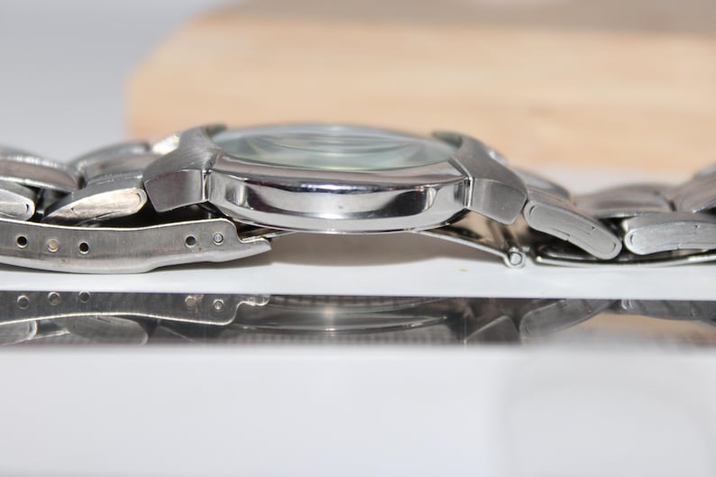 May include: A silver-toned wristwatch with a metallic band. The watch face is visible, and the band is connected with a series of small, linked segments. The watch is displayed on a reflective surface.