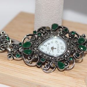 May include: Silver-toned watch with a pear-shaped face and a white dial. The watch features green and clear gemstones and is displayed on a wooden surface. The watch band has an ornate design with additional gemstones.