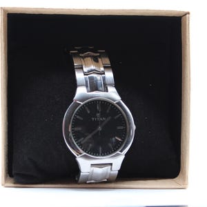 May include: A silver-toned Titan wristwatch with a black dial, presented in a box with a black interior. The watch features a metallic band and is set against a neutral background.