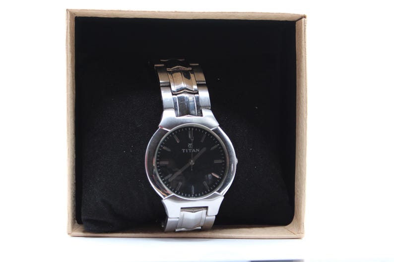 May include: A silver-toned Titan wristwatch with a black dial, presented in a box with a black interior. The watch features a metallic band and is set against a neutral background.