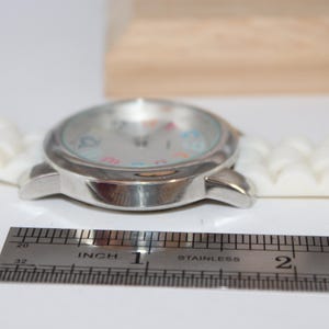 May include: A silver wristwatch with a white silicone band. The watch face has colorful numbers and hands. A stainless steel ruler is next to the watch, showing a measurement of approximately 1 inch.