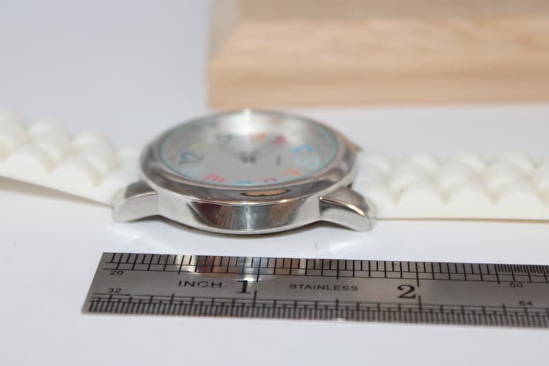 May include: A silver wristwatch with a white silicone band. The watch face has colorful numbers and hands. A stainless steel ruler is next to the watch, showing a measurement of approximately 1 inch.