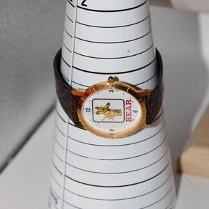 May include: A gold-toned wristwatch with a brown leather band, featuring a white face with a cartoon bear graphic and the word "BEAR". The watch is displayed on a white, cone-shaped bracelet sizing tool.