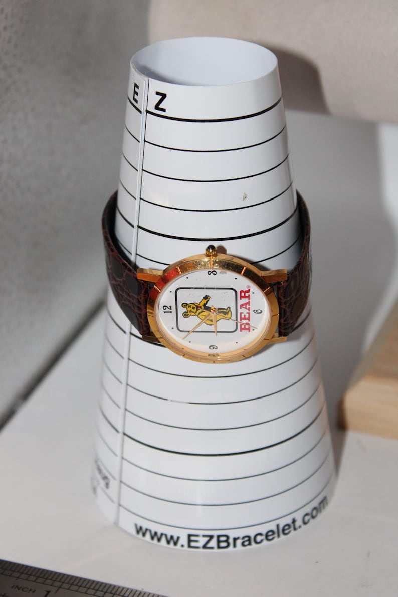 May include: A gold-toned wristwatch with a brown leather band, featuring a white face with a cartoon bear graphic and the word "BEAR". The watch is displayed on a white, cone-shaped bracelet sizing tool.
