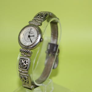 May include: Silver-toned women's watch with a white face, black hands, and Roman numeral hour markers. The watch band features ornate scrollwork and a hinged clasp. The watch is displayed on a clear stand against a light green background.