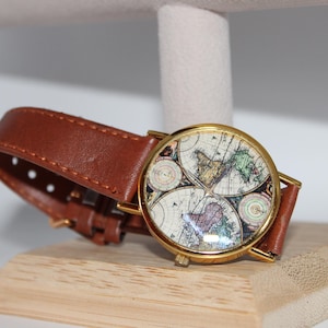 May include: A round wristwatch with a gold-colored case and a brown leather band. The watch face features a vintage map design with various colors. The watch is displayed on a wooden stand.