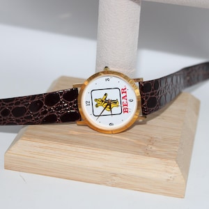 May include: Gold-tone wristwatch with a white face featuring a yellow bear graphic and the word "BEAR" in red. The watch has a brown, textured leather band and a gold buckle. The watch is displayed on a wooden stand.