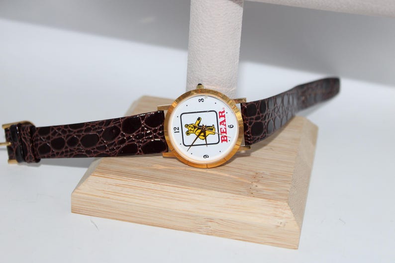 May include: Gold-tone wristwatch with a white face featuring a yellow bear graphic and the word "BEAR" in red. The watch has a brown, textured leather band and a gold buckle. The watch is displayed on a wooden stand.