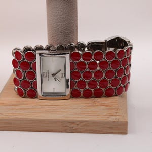 May include: A silver-toned watch with a rectangular face and a band of red, circular accents. The watch is displayed on a wooden block. The watch face has black hands and silver hour markers.
