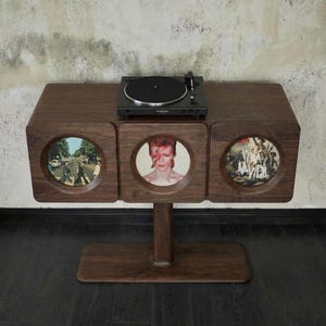 May include: A dark brown wooden record player stand with a black turntable on top. The stand has three circular cutouts displaying album art. The stand is supported by a single, central leg.