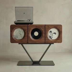 May include: A mid-century modern record player console with a turntable on top. The wooden cabinet features three square compartments, one with a record, one with a framed album cover, and one with a framed illustration. The console is supported by a black metal base.