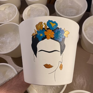 Puede incluir: Maceta de cerámica blanca con un diseño pintado a mano del rostro de Frida Kahlo con una corona floral. Las flores están pintadas en tonos de azul, naranja y dorado.