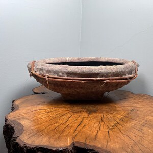Peut inclure: Un bol ovale texturé fait à la main avec un intérieur sombre. Le bol est de couleur gris-brun clair, avec une bande tissée de couleur naturelle autour du bord. Il repose sur une surface en bois rustique.