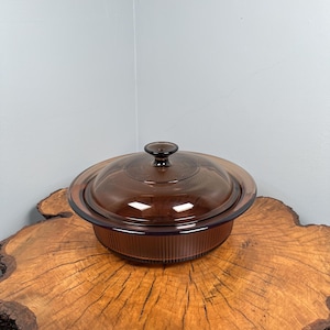 Puede incluir: Un plato para hornear de vidrio marrón con tapa. El plato tiene un borde acanalado y una perilla en la tapa.