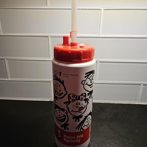 May include: White plastic water bottle with a red lid and a straw. The bottle has a cartoon design of people's faces and the text "Poulet Frit Kentucky".