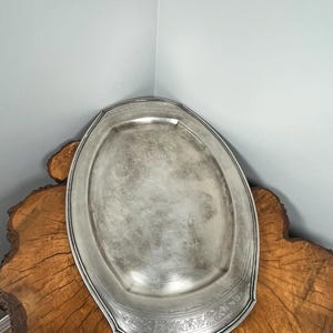 May include: A silver oval serving tray with an ornate design around the edge. The tray is sitting on a wooden surface.