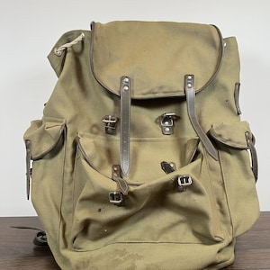 May include: Olive green canvas backpack with brown leather straps and accents. Features multiple pockets, metal buckles, and a top flap closure. The backpack has a vintage, worn appearance.