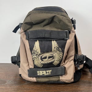May include: A brown and black backpack with a winged emblem and the word "SPLIT". The backpack has adjustable straps and multiple compartments. The main body is a mix of brown and black panels. The emblem is gold-coloured.
