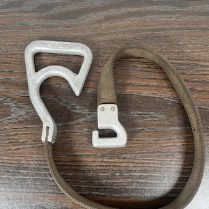 Könnte beinhalten: Ein Vintage-Metallgriff mit dem Wort "AKRON" und einem passenden Haken, der mit einem khaki-farbenen Riemen verbunden ist. Der Griff und der Haken sind silberfarben. Der Riemen besteht aus einem gewebten Material.