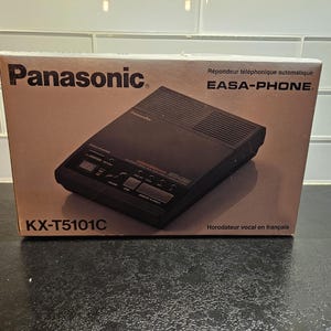 May include: A black Panasonic EASA-PHONE answering machine with a digital display and a built-in speaker. The box features the text "Répondeur téléphonique automatique EASA-PHONE" and "KX-T5101C".