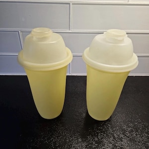 Puede incluir: Dos botellas de plástico amarillo pálido con tapas blancas. Las botellas tienen una base redondeada y una parte superior acampanada. Las tapas tienen una pequeña abertura. Las botellas están sobre una superficie negra.