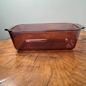 Puede incluir: Una fuente rectangular de vidrio transparente de color marrón rojizo con asas. La fuente está vacía y se encuentra sobre una superficie de madera. El vidrio tiene un ligero tinte y el diseño general es simple y funcional.