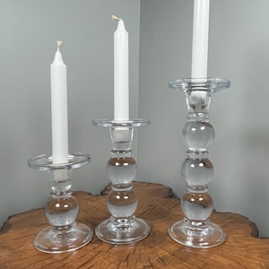 May include: Three clear glass candle holders with white candles. The candle holders have a round base, a stem with spherical accents, and a circular top. The candles are white and taper to a point.