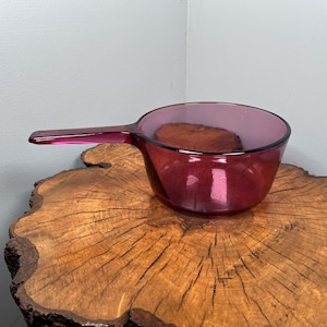 Puede incluir: Una cacerola de vidrio rojo rubí vintage con un asa. La cacerola está sobre una superficie de madera. La cacerola es de un color rojo intenso y transparente, y el asa está sujeta al lateral.