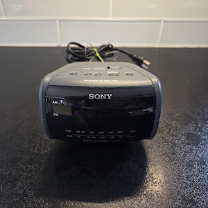 May include: A grey Sony Dream Machine alarm clock with a digital display. The clock has buttons for snooze/sleep off and radio frequency settings. The clock is on a dark surface.
