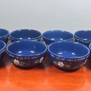Peut inclure: Un ensemble de huit bols en céramique bleu profond disposés sur deux rangées. Chaque bol présente une marguerite blanche peinte à la main avec des feuilles vertes près de la base et du texte français en cursive blanche le long du bord.