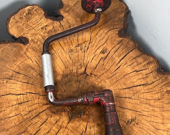 Op de afbeelding: Een antieke handboor met een rood en zwart handvat en een zilveren grip. De boor staat tegen een houten achtergrond met een natuurlijk nerfpatroon. De tool vertoont tekenen van slijtage.