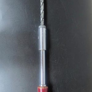 May include: A metal hand drill with a red wooden handle. The drill has a silver shaft with a spiral design and a silver chuck. The handle is a deep red colour with a yellowed base. The drill is set against a dark background.