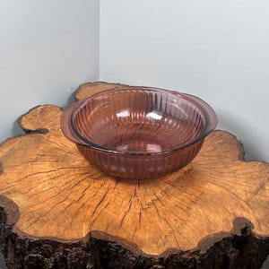 May include: A vintage, round, glass bowl in a muted rose colour. The bowl has a ribbed design and two handles. It sits on a rustic, wooden tree stump with a natural edge. The background is a neutral grey.
