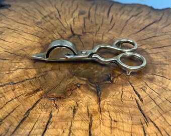 May include: Brass cigar cutter with a scissor-like design. The cutter has a round blade and a handle with a loop.