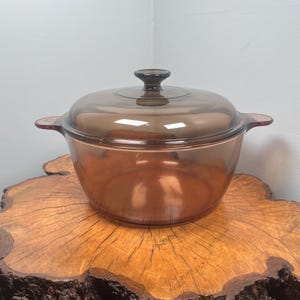 May include: A round, transparent amber-coloured glass casserole dish with a matching glass lid. The pot features two integrated side handles and a central knob on the lid. The glass has a smooth, polished finish and a deep brown tint throughout.