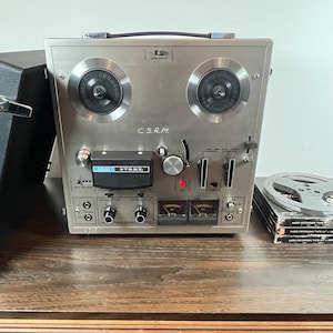 Peut inclure: Un magnétophone à bobines argenté vintage avec un étui de transport noir et une pile de bandes d'enregistrement audio. L'enregistreur comporte deux grandes bobines, des cadrans et le texte "C.S.R.M." et "AKAI 4700DS".