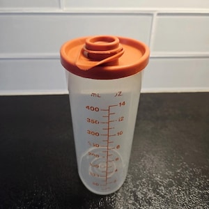 Puede incluir: Un vaso medidor de plástico transparente con tapa naranja. El vaso tiene marcas de medición rojas en mililitros y onzas. La tapa tiene un pequeño pico. Para uso en la cocina.
