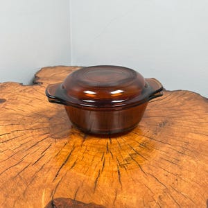 Könnte beinhalten: Eine braune Glas-Auflaufform mit Deckel. Die Form hat zwei Griffe an den Seiten. Die Form steht auf einer Holzoberfläche.